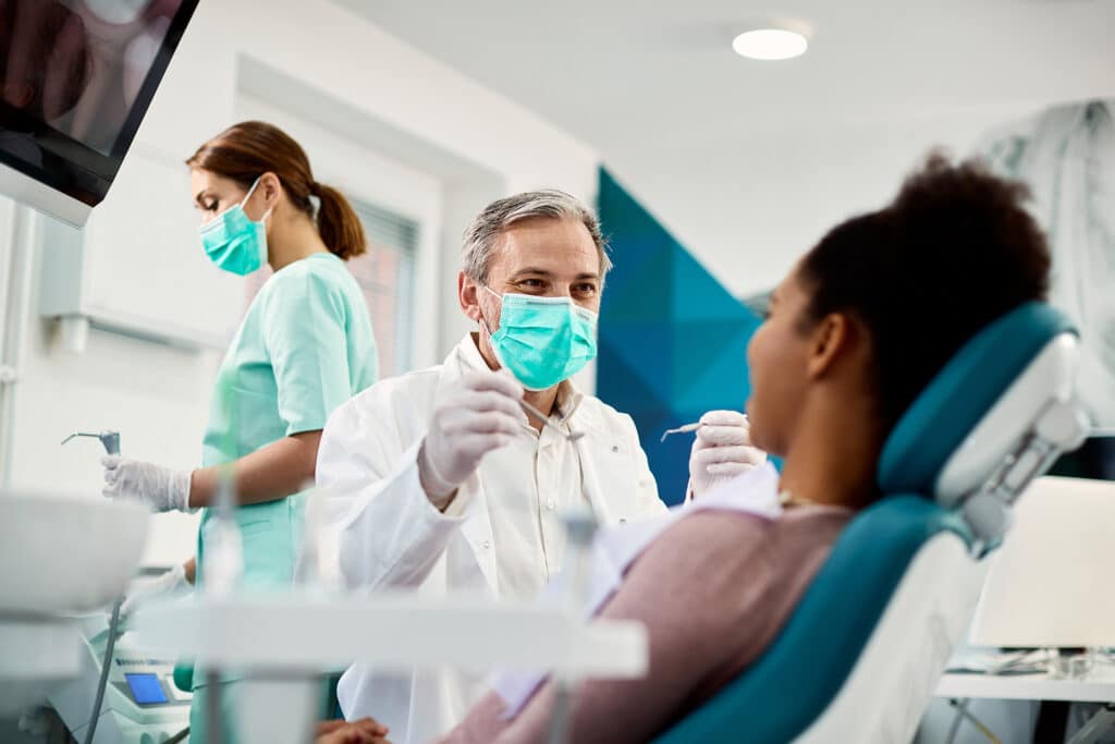 Smiling dentist with face mask talking to Black woman during dental procedure at dental clinic.